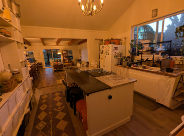 Cozy kitchen with white cabinets, granite island, and open shelves. Warm lighting, plants by the window, leads into a spacious living room with rustic wooden floors.