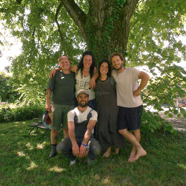 Fünf junge Leute stehen an einem sonnigen Tag fröhlich unter einem großen, grünen Baum. Vier von ihnen stehen Arm in Arm, während einer davor hockt, und alle lächeln herzlich.