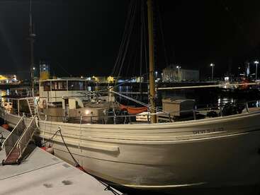 Un barco clásico de madera está atracado en un puerto deportivo por la noche, iluminado por suaves luces. El agua tranquila refleja las luces de los edificios cercanos.