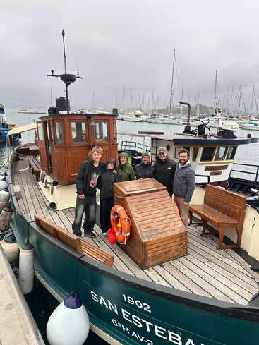 Six people stand on the deck of a classic wooden boat named "San Esteban," docked at a marina on a cloudy, overcast day. Sailboats nearby.