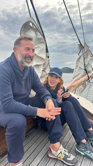 A bearded man and a young woman sit on a sailboat, both dressed warmly, smiling and talking, with sails and cloudy skies in the background.