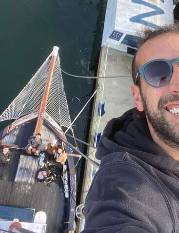 Un hombre con gafas de sol se hace un selfie desde lo alto del mástil de un barco. Abajo, varias personas en la cubierta miran hacia arriba, atracadas junto al agua.
