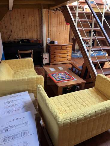 Esta acogedora habitación cuenta con dos sofás amarillos, muebles de madera, una escalera, papeles de planos de barcos, un juego de mesa, una guitarra y suaves paneles de madera en las paredes.