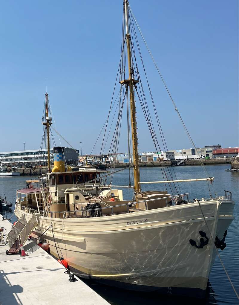 A cream-colored vintage boat with tall masts is docked at a harbor under a clear blue sky, surrounded by industrial buildings and calm water.