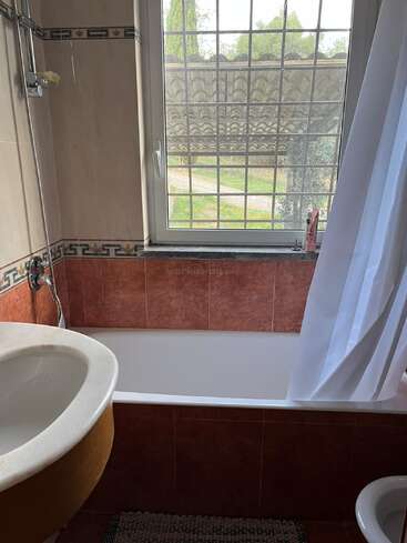 Dieses Bild zeigt ein kleines Badezimmer mit einer Badewanne, einer Dusche, einem Waschbecken und einem Fenster mit Gittern. Natürliches Licht fällt ein, und draußen sind Bäume zu sehen.