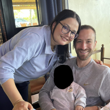 Ein lächelndes Paar posiert zusammen in einem Haus, mit einem verschwommenen Kindergesicht auf dem Schoß des Mannes. Sie wirken glücklich und genießen einen gemütlichen Familienmoment zu Hause.
