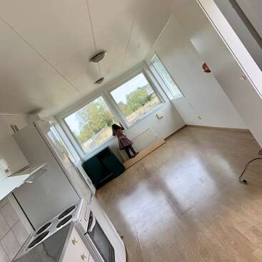 Una niña pequeña con un vestido está de pie junto a unos grandes ventanales en una habitación vacía e iluminada por el sol, con una zona de cocina, suelos de madera y una perspectiva inclinada.