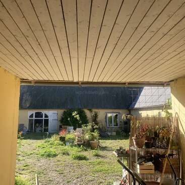Esta imagen muestra un acogedor patio con plantas en macetas, muebles de exterior y herramientas de jardinería bajo un tejado de madera, frente a una casa y un jardín rústicos de color amarillo.