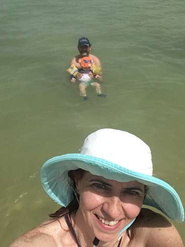 A imagem mostra uma mulher usando um chapéu de sol tirando uma selfie com um homem e uma criança na água atrás dela.