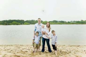 Das Bild zeigt eine fünfköpfige Familie, die an einem Sandstrand steht, mit einem ruhigen Gewässer und einer üppigen grünen Landschaft im Hintergrund.