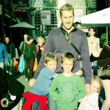 Das Bild zeigt einen Mann, der mit zwei Kindern vor einer belebten Straße posiert, mit einer Menschenmenge und Regenschirmen im Hintergrund.
