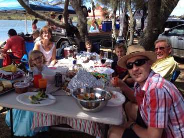 Das Bild zeigt eine Familie, die eine Mahlzeit im Freien genießt. Sie sitzt an einem Tisch mit Essen und Getränken, umgeben von einem ruhigen See und Bäumen.