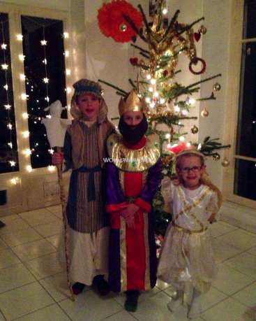 Das Bild zeigt drei kostümierte Kinder, die vor einem Weihnachtsbaum mit festlicher Atmosphäre und blinkenden Lichtern posieren.