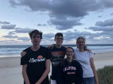Vier Menschen stehen bei Sonnenuntergang an einem Strand und lächeln. Der Himmel ist voller dramatischer Wolken und die Wellen des Meeres rollen hinter ihnen her.