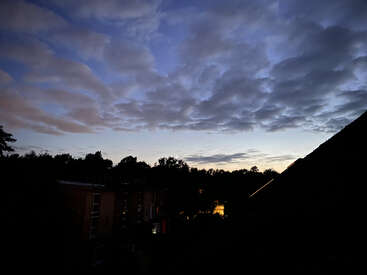 The image captures a serene evening sky with scattered dark clouds, soft blue hues, silhouettes of trees, buildings, and faint streetlights glowing peacefully below.