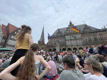 A large crowd gathers in a historic town square, with people sitting and standing. German and EU flags are displayed on the ornate building in the background.