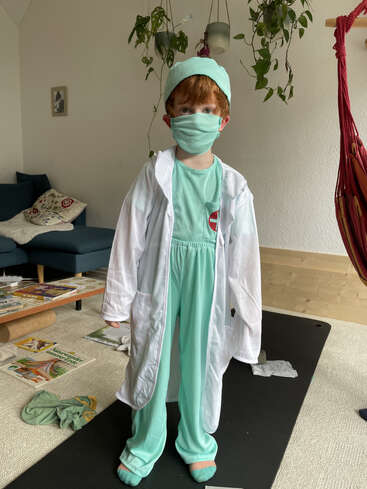 A young child dressed as a doctor stands indoors, wearing green scrubs, a face mask, and a white coat. Plants hang above; books are scattered nearby.