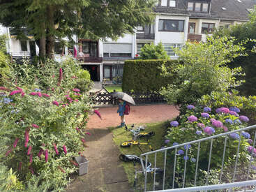 A child with a backpack and umbrella stands on a garden path. Scooters and bikes are scattered, surrounded by blooming flowers and green bushes. Urban homes behind.