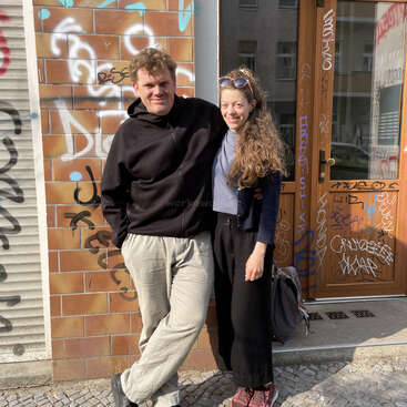 A smiling man and woman stand close together outside, in front of a brick wall and door covered with colorful graffiti, enjoying a sunny day.