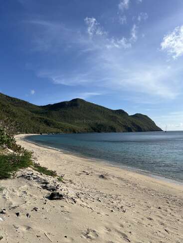 A serene sandy beach meets clear blue water, with lush green hills under a bright sky. Calm, peaceful scenery, perfect for relaxation and unwinding in nature.