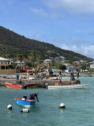 A small harbor scene with colorful boats, people working on the dock, turquoise water, mountains in the background, palm trees, and houses under a bright blue sky.