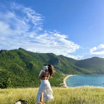 A woman in a white skirt stands on a grassy hill, holding a dog. Beautiful green mountains, blue sky, and a serene bay create a stunning backdrop.