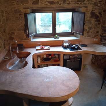Essa cozinha rústica tem paredes de pedra, bancadas de madeira curvas, uma pia única, prateleiras abertas com pratos, fogão, forno, janelas abertas e iluminação natural que dá um clima aconchegante.