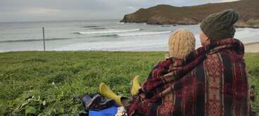 Duas pessoas, bem aconchegadas em um cobertor estampado, sentam-se em uma colina gramada com vista para os surfistas pegando ondas no mar perto de um litoral rochoso e pitoresco.