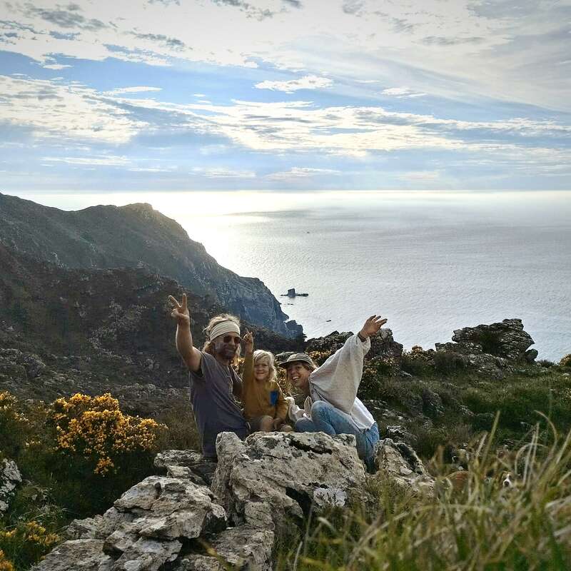A imagem mostra três indivíduos posando em um afloramento rochoso com vista para um vasto corpo de água, tendo como pano de fundo montanhas e um céu nublado.