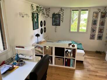 Um quarto aconchegante com paredes brancas e piso de madeira, com uma cama, uma mesa organizada, estantes, quadros na parede, trepadeiras pendentes e luz natural que entra pelas janelas grandes.