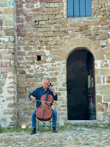 Um homem está sentado num banquinho, tocando violoncelo em frente a um prédio antigo de pedra com uma porta em arco e janelas pequenas, criando um ambiente rústico.