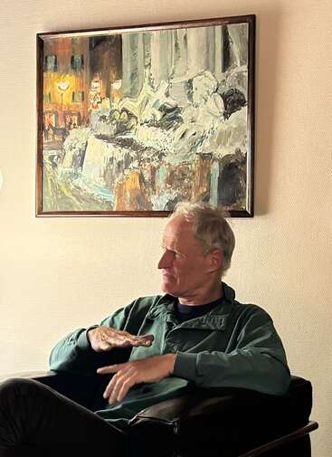 Um homem de cabelos grisalhos está sentado numa cadeira, gesticulando enquanto fala. Atrás dele, há um quadro colorido retratando uma paisagem urbana com formas abstratas.