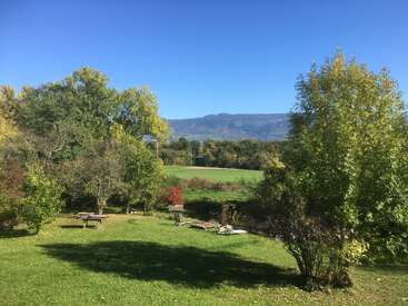 Eine sonnige Landschaft mit grünem Gras, vereinzelten Bäumen, fernen Hügeln und ein paar Holzbänken und Liegestühlen. Der Himmel ist klar und strahlend blau.
