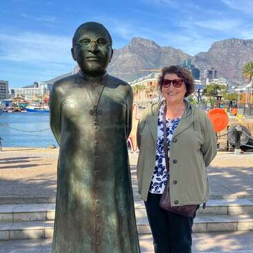 Una mujer sonriente posa junto a una estatua de bronce de un hombre con gafas y una cruz, con las montañas y el paisaje urbano visibles en el fondo soleado.
