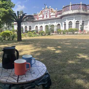 A small table with three colorful cups and a black kettle sits on grass in a garden, with a grand white historical building in the background.