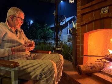 An elderly man sits comfortably by a glowing fireplace at night, holding a drink, enjoying warmth and solitude, with a garden and house visible outside.