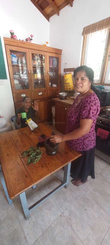 Two women are in a cozy kitchen. One is grinding leaves with a mortar and pestle, while the other sits, smiling and holding a green cup.