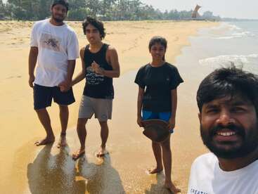 Four friends are standing and smiling on a sunny beach, with gentle waves nearby. They appear happy, enjoying their time together on the sandy coastline.
