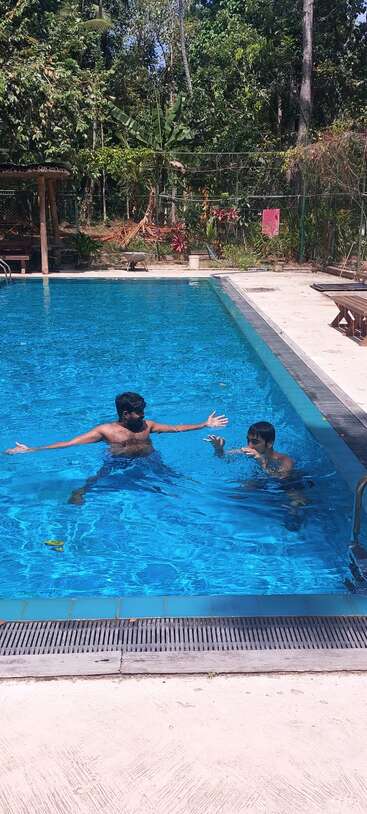 Two people are enjoying a sunny day swimming in a clear blue pool surrounded by lush green trees and nature, creating a relaxing tropical atmosphere.