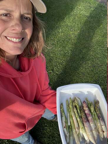 Una persona sonriente, vestida con una sudadera roja con capucha y una gorra beige, sostiene una bandeja de espárragos recién cosechados mientras está de pie sobre la hierba verde, disfrutando de un día soleado.