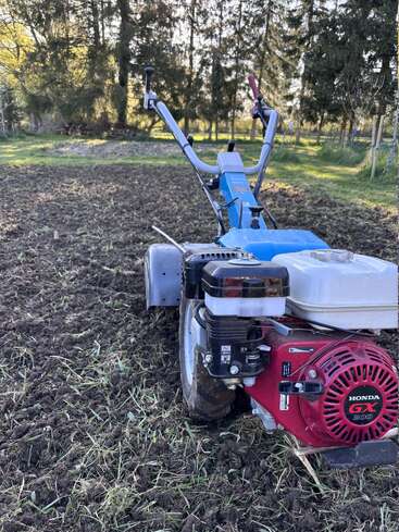 Una motocultivadora con motor Honda GX200 está aparcada sobre tierra recién labrada en un jardín, rodeada de césped verde y árboles altos.