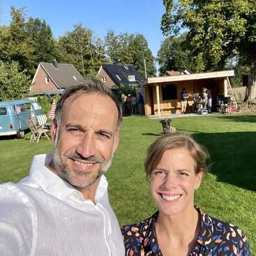 A man and woman smile for a selfie outdoors, with a sunny backyard, green grass, wooden shed, blue van, and other people in the background.