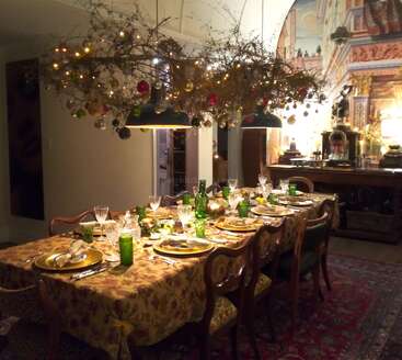 Une salle à manger festive présente une longue table garnie de plats et de verres élégants, entourée de chaises anciennes, sous des branches suspendues décorées de boules et de lumières.