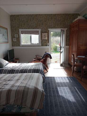 Cette chambre confortable dispose de deux lits simples, d'une télévision, d'un fauteuil à bascule, d'une grande armoire, d'un bureau, d'un papier peint à motifs, de la lumière du soleil qui entre par une porte ouverte et d'un jardin luxuriant à l'extérieur.