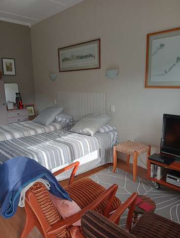 Une chambre confortable et ensoleillée comprend deux lits jumeaux rayés, une chaise en bois avec des couvertures, une télévision, des petits meubles, des œuvres d'art encadrées et des murs doux aux couleurs neutres.