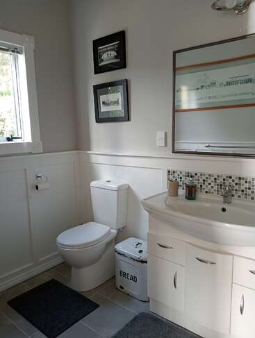 Il s'agit d'une salle de bains moderne et lumineuse avec des murs blancs, des toilettes, un lavabo, un miroir, des œuvres d'art murales, une fenêtre, des tapis gris et une corbeille à pain inhabituelle à côté des toilettes.