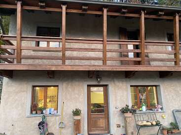 Dieses Bild zeigt ein rustikales Haus mit einem Holzbalkon, zwei Fenstern, einer Holztür, Pflanzen, Reinigungsutensilien und einer Bank draußen, was eine gemütliche Atmosphäre schafft.