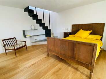 In diesem Zimmer gibt es ein Holzbett mit leuchtend gelber Bettwäsche, eine moderne schwarze Treppe, einen Holzstuhl, Wandregale, eine Vase und einen Holzboden. Minimalistisches Design.