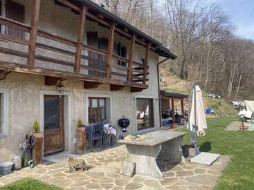 Ein rustikales zweistöckiges Haus mit einem Holzbalkon, einer Steinterrasse, einem Tisch im Freien, einem Grill, gestapelten Stühlen, einem Sonnenschirm und einer Katze, die draußen auf dem Boden herumläuft.