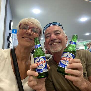 Ein lächelndes älteres Paar genießt gemeinsam Seybrew-Biere, hält die Flaschen für ein Selfie in einem hellen Innenraum hoch und wirkt während seines Ausflugs glücklich und entspannt.
